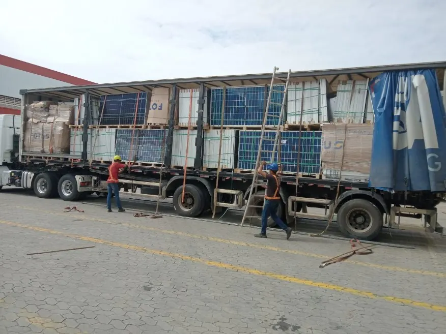 Carreta sider sendo carregada com paletes de módulos fotovoltaicos em um pátio, com dois operadores de capacete e colete fixando a carga