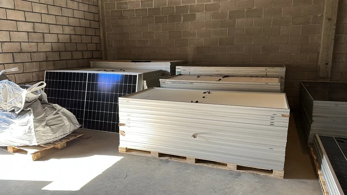 Módulos fotovoltaicos empilhados sobre paletes de madeira dentro de um galpão coberto, com a face de vidro para baixo e um big bag ao lado