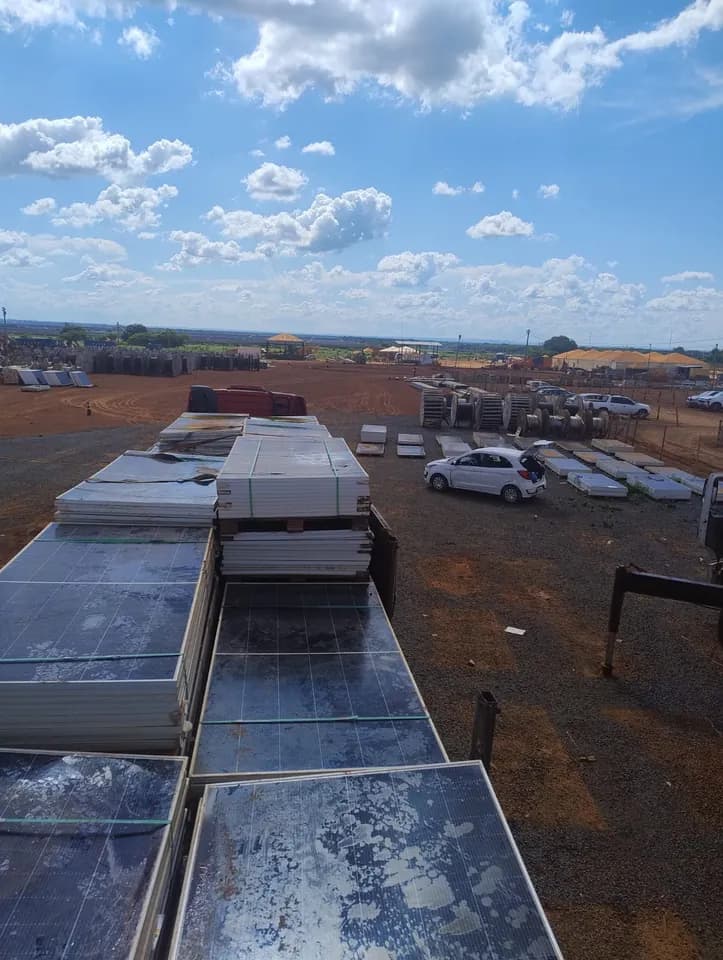 Vista do alto de um caminhão aberto carregado com módulos fotovoltaicos empilhados, alguns com o vidro manchado e trincado, num canteiro de terra com carros e outras pilhas de módulos ao fundo