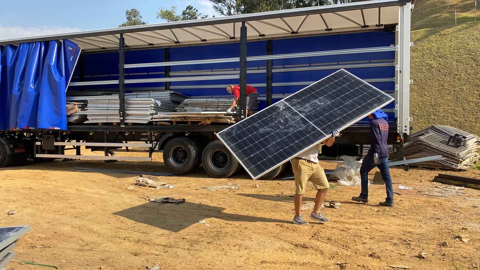 Dois operadores carregam um módulo fotovoltaico com o vidro trincado até uma carreta sider de lona azul aberta, estacionada em terreno de terra, com outros módulos já empilhados dentro