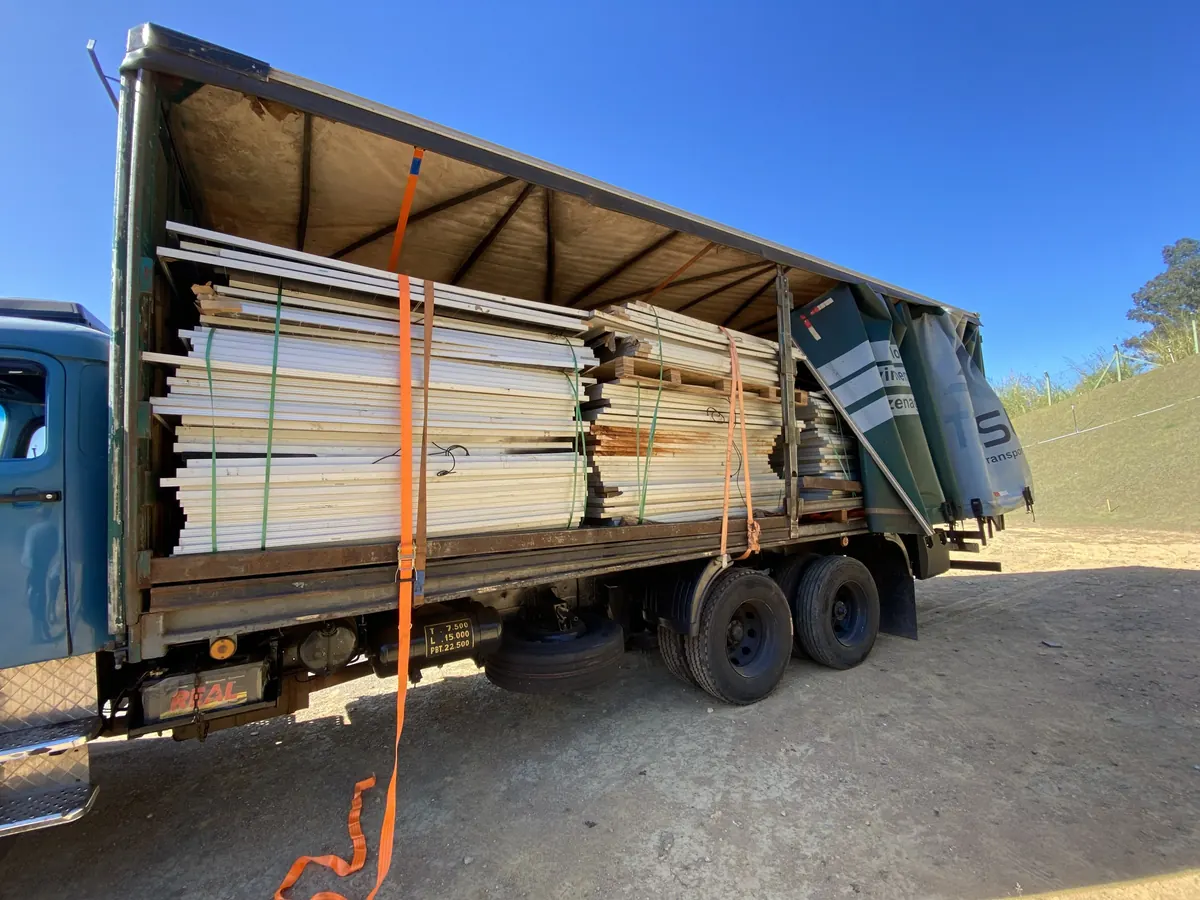 Truck with its side tarp open, loaded with photovoltaic modules stacked in batches on pallets and secured with straps, parked on a dirt yard