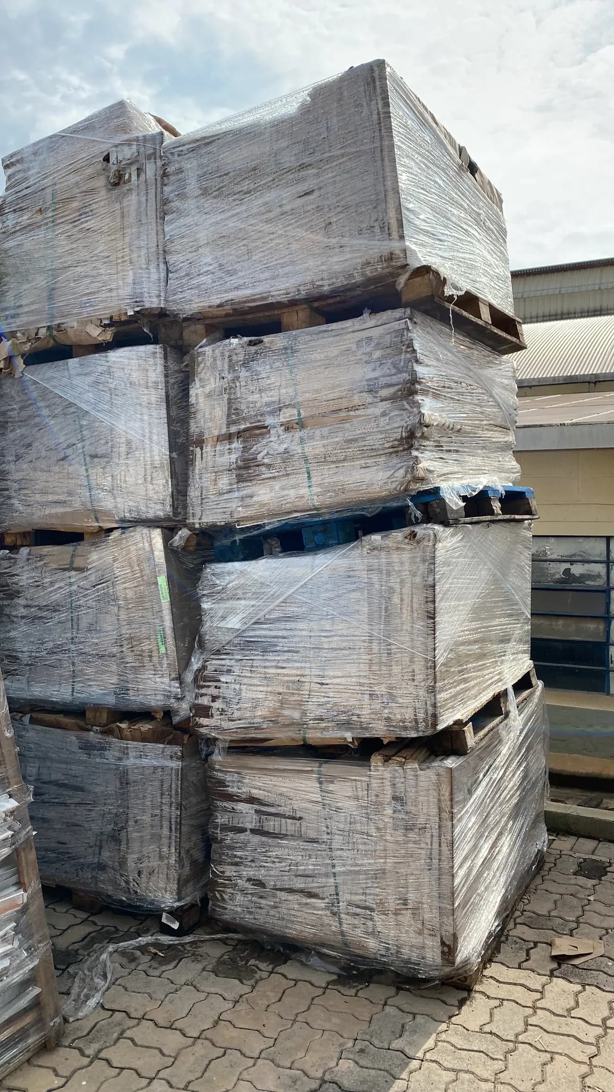 Pilha alta de paletes de módulos fotovoltaicos embalados em filme plástico, empilhados em quatro níveis sobre piso de bloquetes, ao lado de um galpão