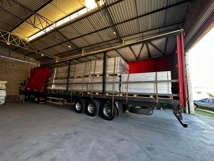 Carreta de lona vermelha com a lateral aberta dentro de um galpão coberto, carregada com módulos fotovoltaicos empilhados sobre paletes