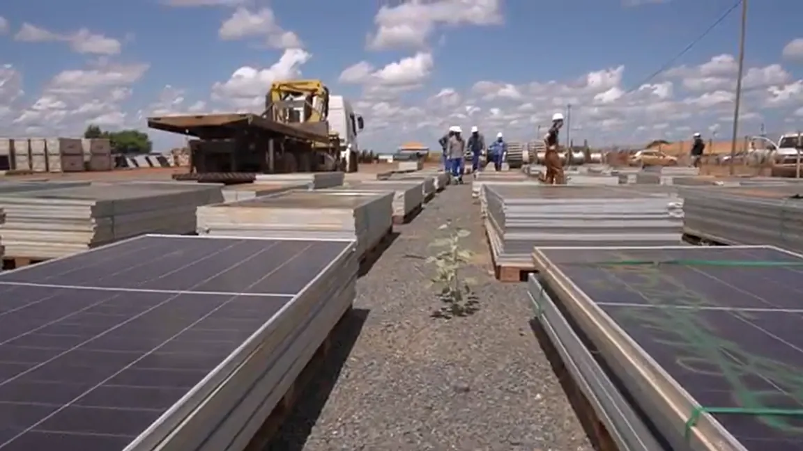 Paletes de módulos fotovoltaicos alinhados no pátio de uma usina, com equipe de capacete ao fundo e caminhão com guindaste pronto para a coleta