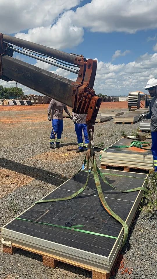 Equipe com uniforme e capacete prende com cintas um palete de módulos danificados ao braço de uma máquina para içamento no pátio da usina
