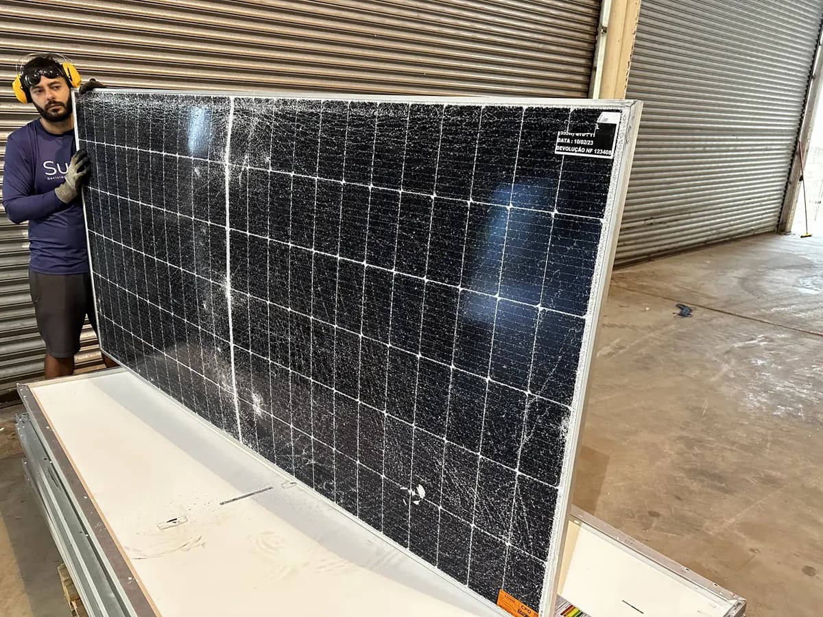Operador com protetor auricular e luvas segurando em pé um módulo fotovoltaico com trincas espalhadas por todo o vidro, dentro de um galpão