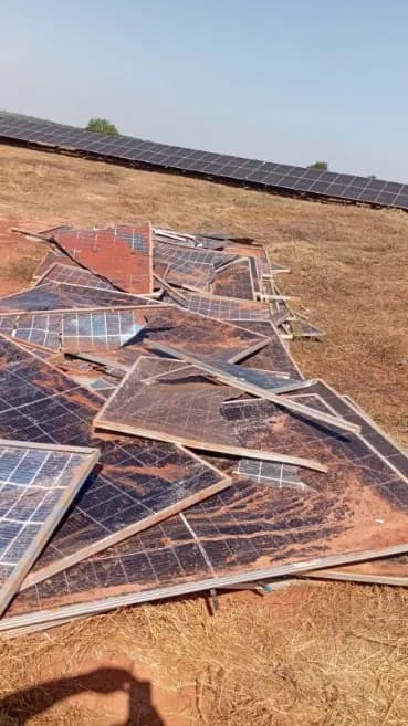 Módulos fotovoltaicos quebrados e retorcidos espalhados no solo de uma usina solar, com fileiras de painéis instalados ao fundo