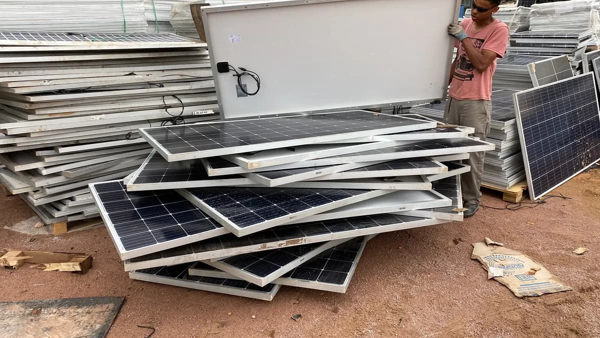 Pilha de painéis solares danificados sobre chão de terra vermelha, com um operador de luvas movendo um módulo pela parte traseira e lotes empilhados ao fundo