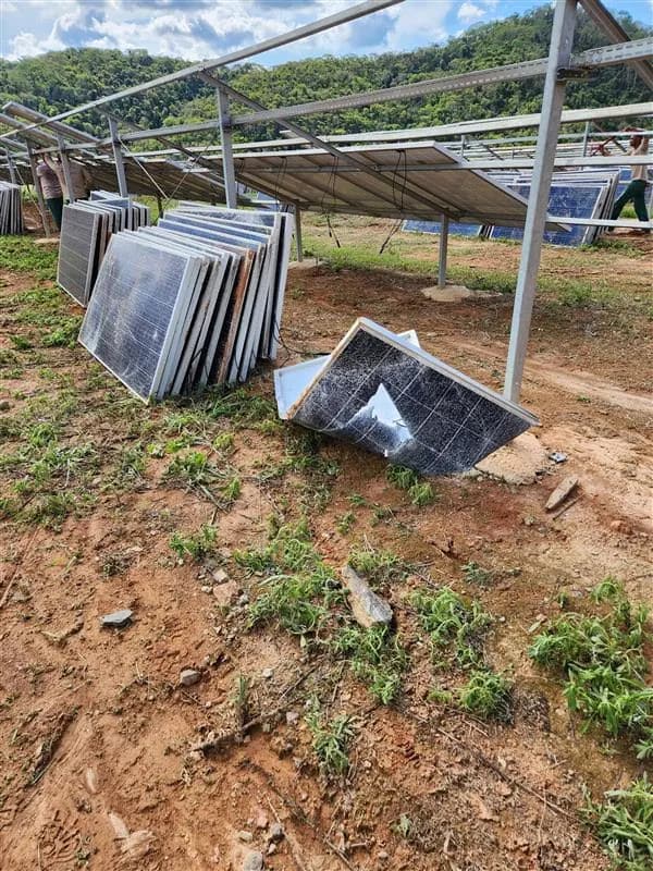 Módulos fotovoltaicos danificados encostados uns nos outros sobre o solo de terra vermelha de uma usina, um deles com o vidro estilhaçado, sob a estrutura metálica de fixação