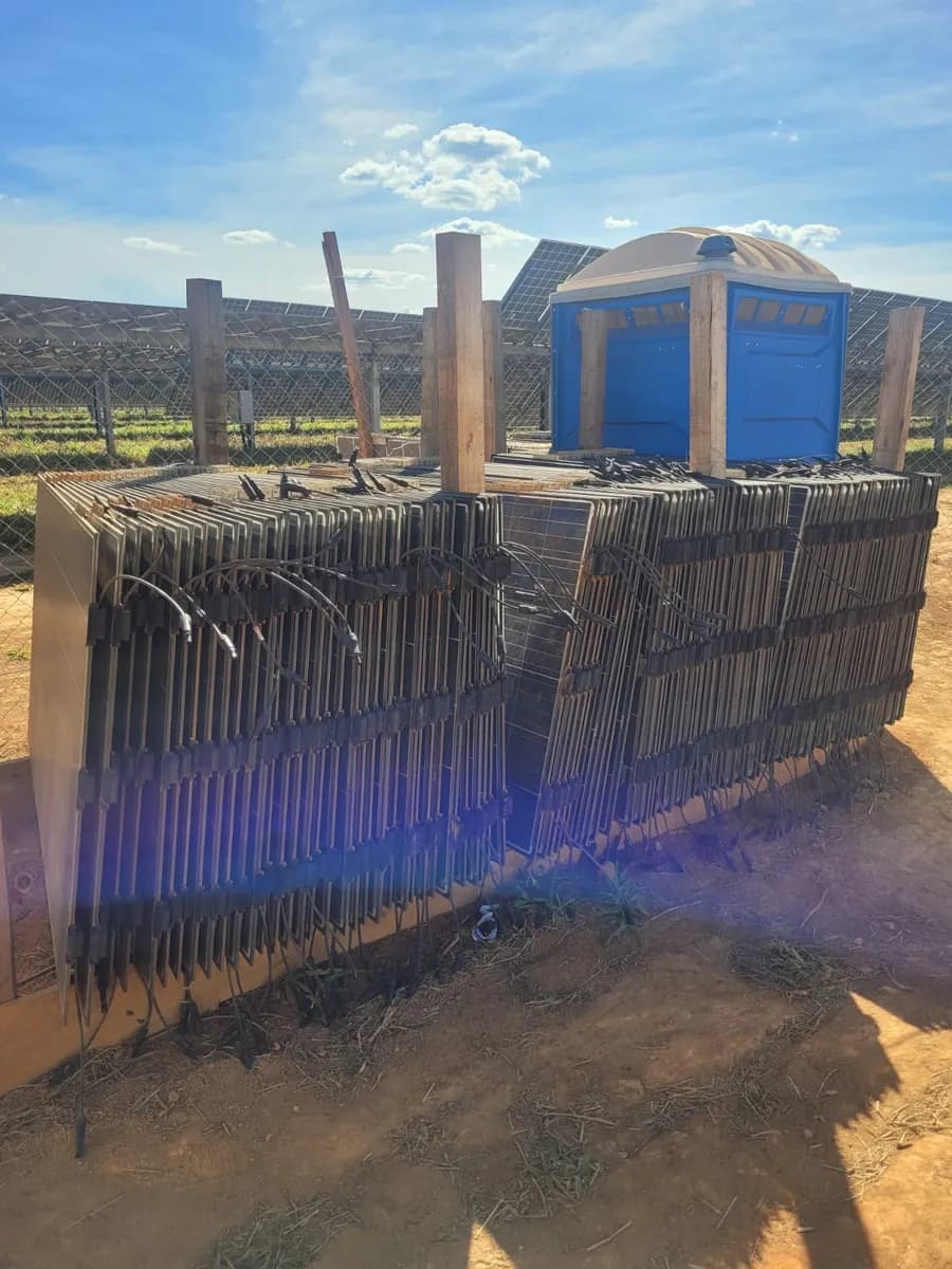 Molduras de alumínio de módulos fotovoltaicos empilhadas na vertical em um pátio de terra de usina, com estacas de madeira e um banheiro químico azul ao fundo