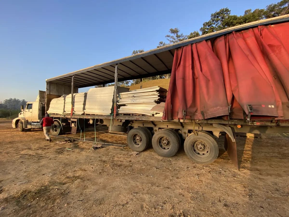 Carreta com lona vermelha aberta em pátio de terra, carregada com pilhas de módulos fotovoltaicos retirados de campo, com um operador ao lado da cabine