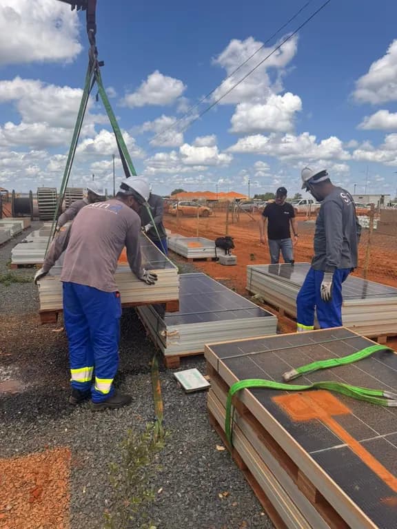 Equipe com uniforme da SunR e capacete organiza módulos fotovoltaicos empilhados em paletes de madeira, com cinta de içamento presa a um lote, em pátio de terra vermelha