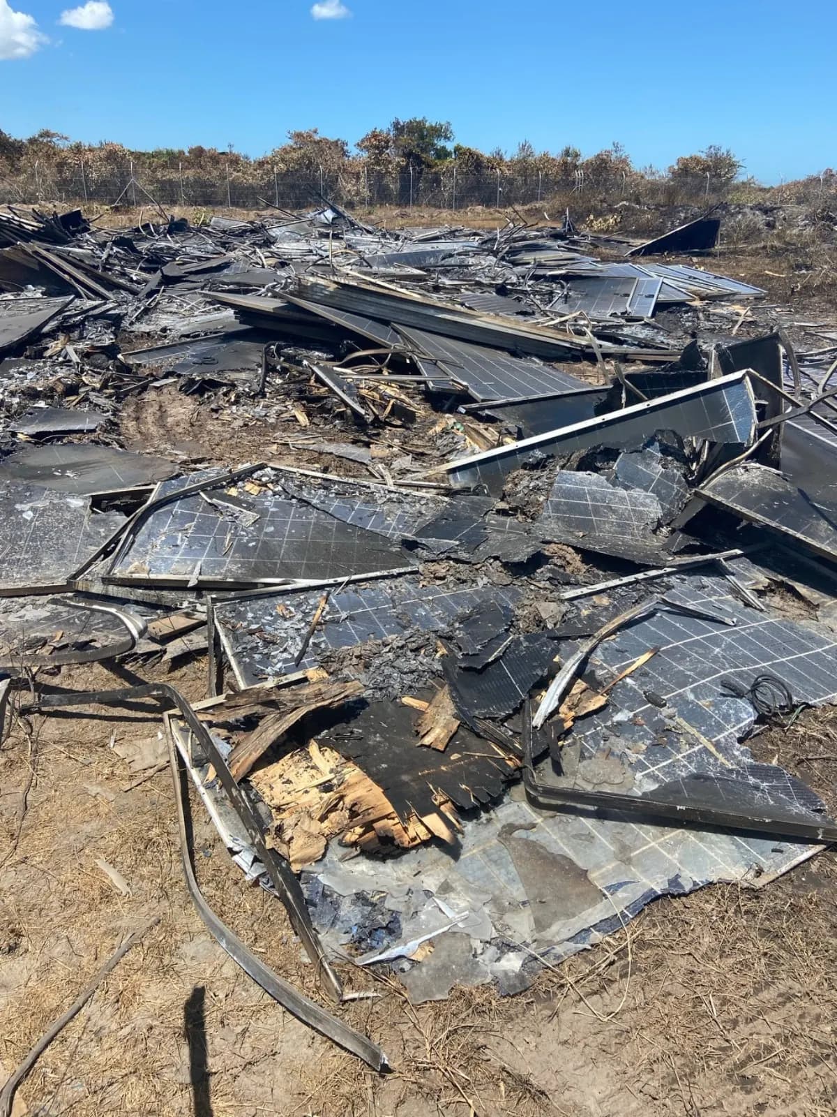 Restos de módulos fotovoltaicos queimados, com vidro estilhaçado, molduras retorcidas e madeira carbonizada, espalhados no chão de uma usina após incêndio